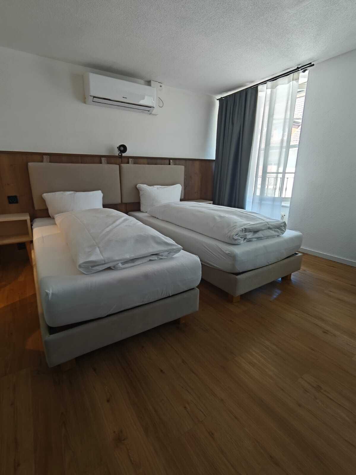 Une chambre d’hôtel avec deux lits, un plancher en bois, une fenêtre avec des rideaux et un climatiseur fixé au mur.