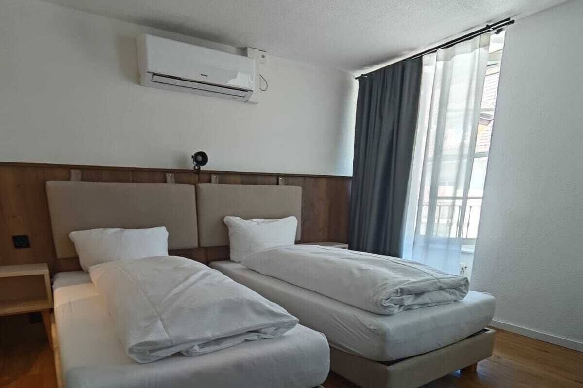 Ein Hotelzimmer mit zwei Betten, Holzboden, einem Vorhangfenster und einer Klimaanlage an der Wand.