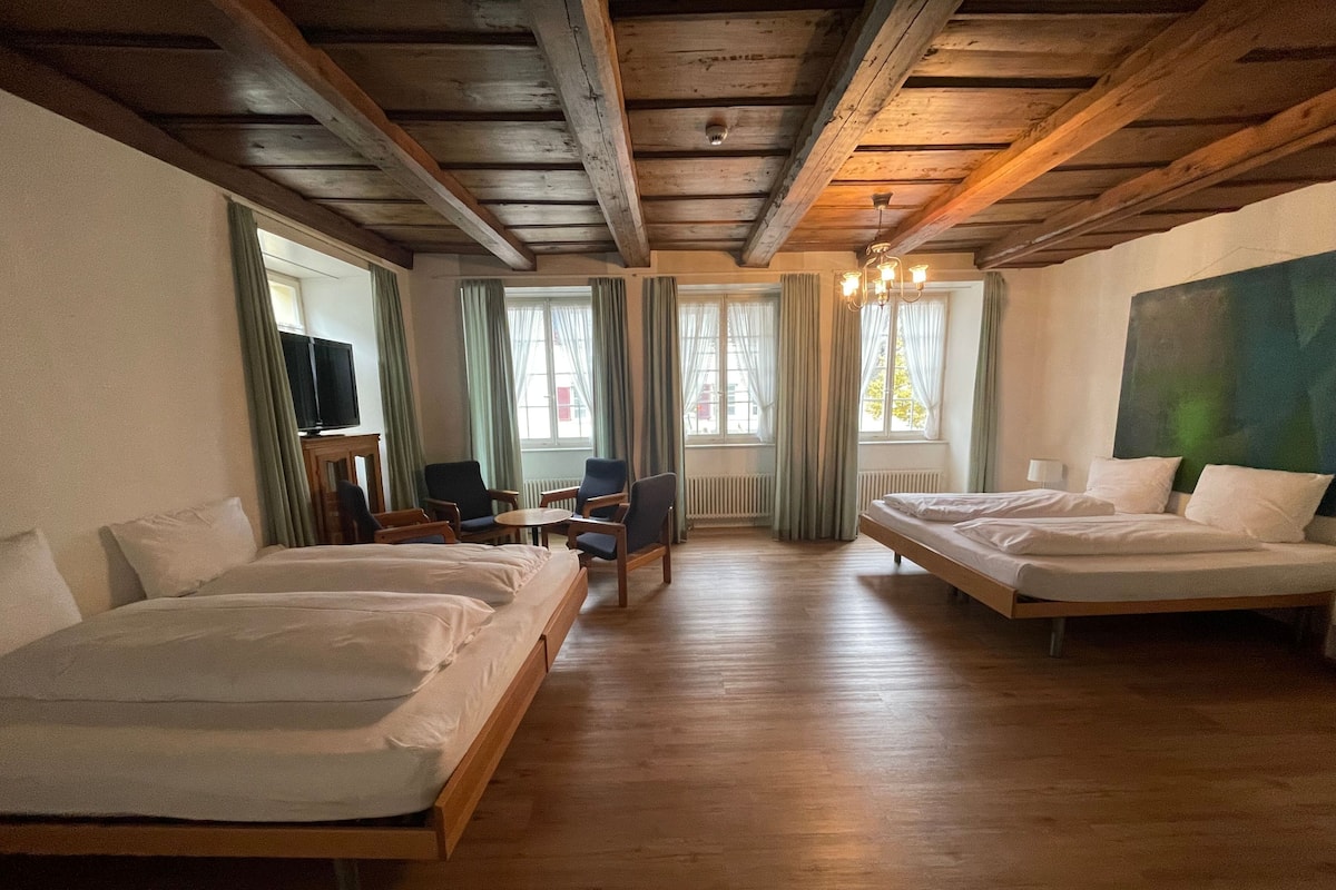 Ein Hotelzimmer mit zwei Einzelbetten, einem kleinen Tisch, zwei Sesseln, einem Fernseher und einem großen Gemälde an der Wand.