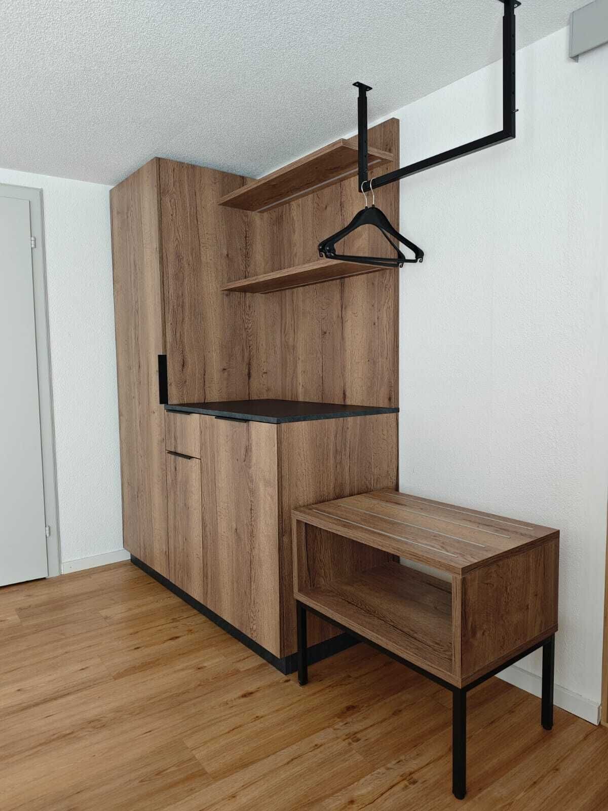 A room with a wooden wardrobe, a black metal shelf, and a wooden table.