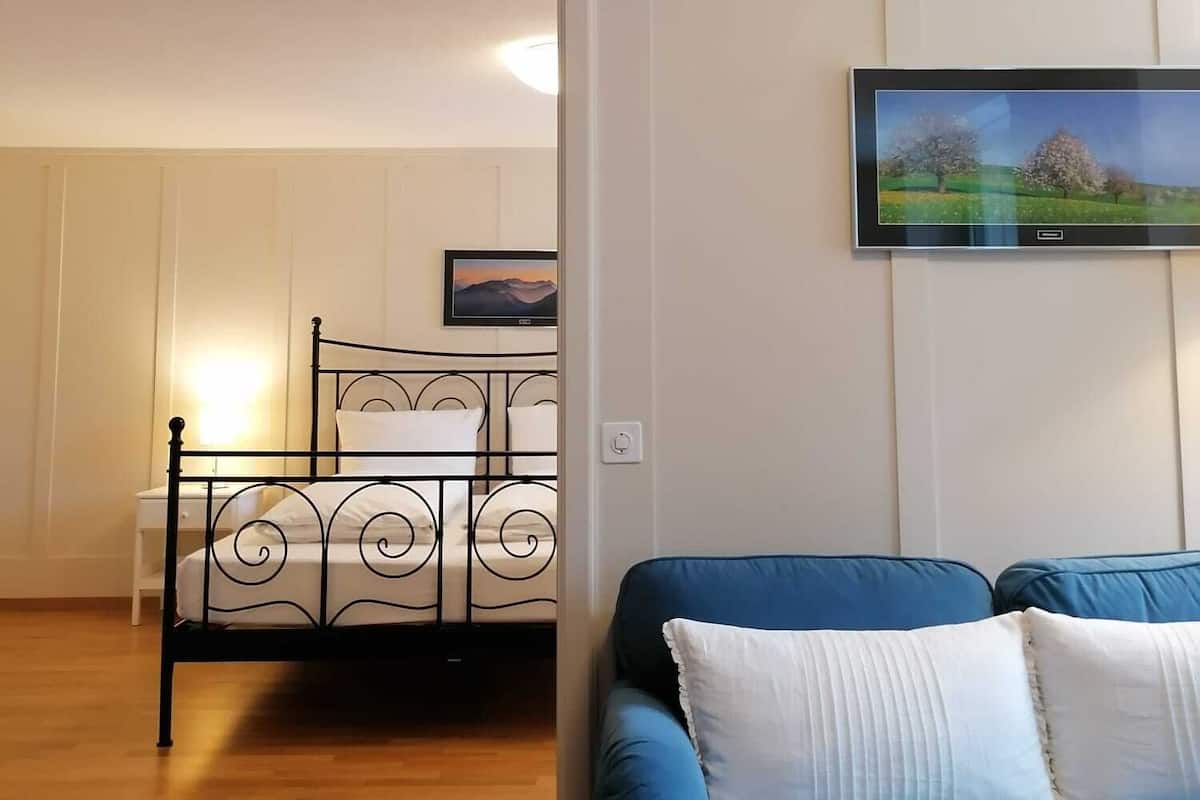 Ein Zimmer mit einem blauen Sofa, einem Metallbettgestell, einem kleinen Nachttisch und einem an der Wand montierten Fernseher.
