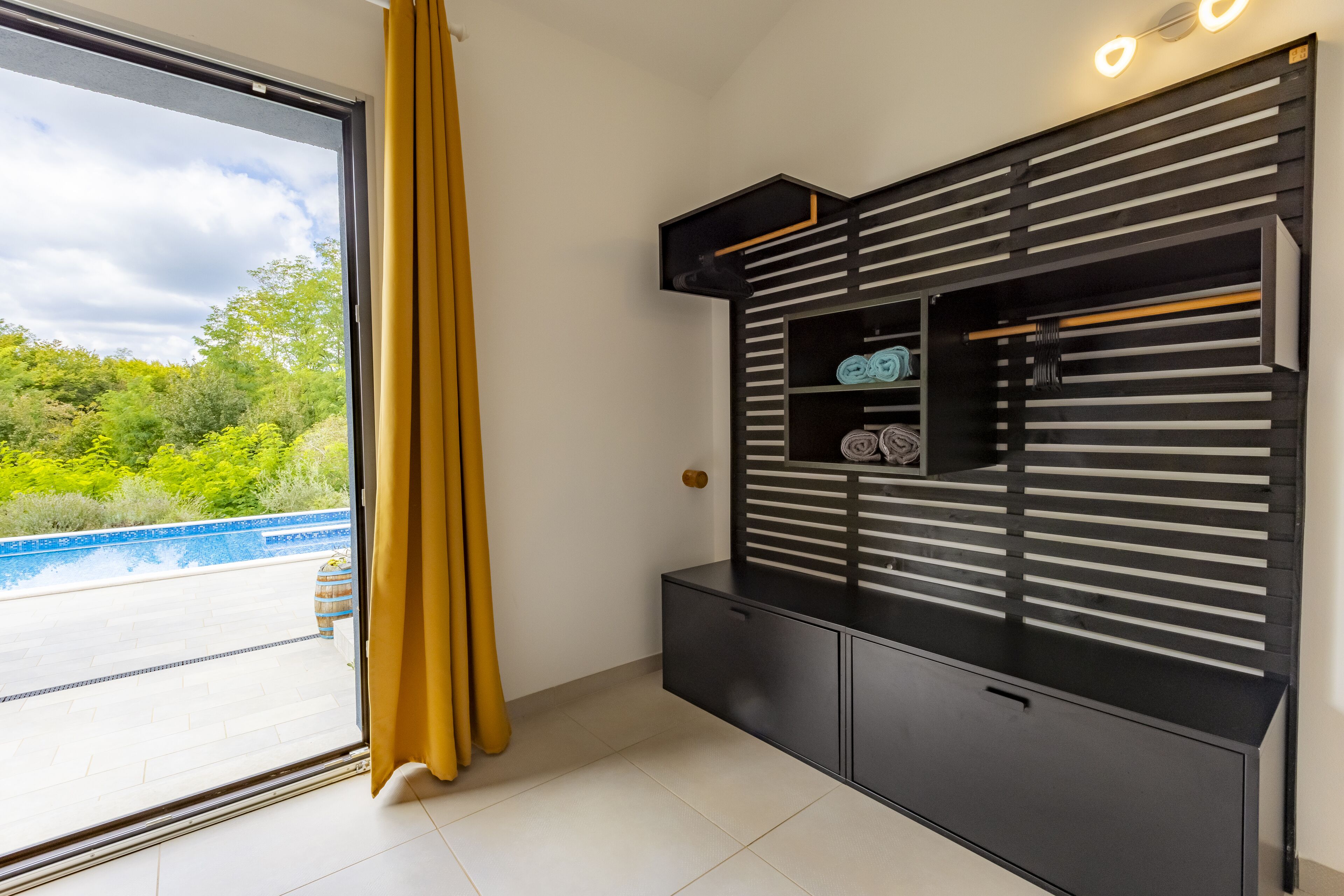 Une pièce dotée d’une grande porte-fenêtre coulissante donnant sur une piscine, d’un meuble de rangement noir avec des étagères et d’un rideau jaune.