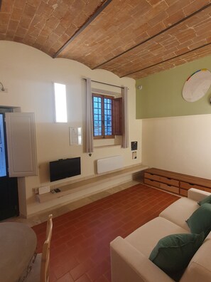 Una sala de estar con un sofá blanco, un televisor sobre un mueble bajo, una ventana con cortinas, un radiador y un reloj de pared.
