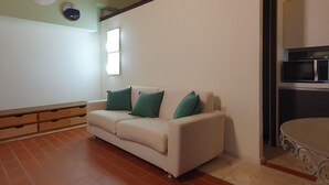 Una sala de estar con un sofá blanco, cojines verdes, un mueble de madera, un adorno circular en la pared y una zona de cocina con un microondas.