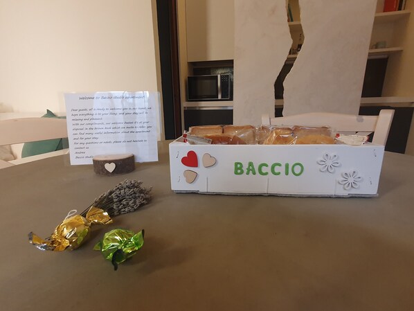 Una cesta de bienvenida con una nota, una caja con el rótulo 'BACCIO' que contiene pan y bombones envueltos sobre una mesa.