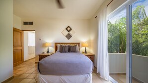 A bedroom with a large bed, two bedside tables with lamps, a ceiling fan, and a view of greenery through a sliding glass door.