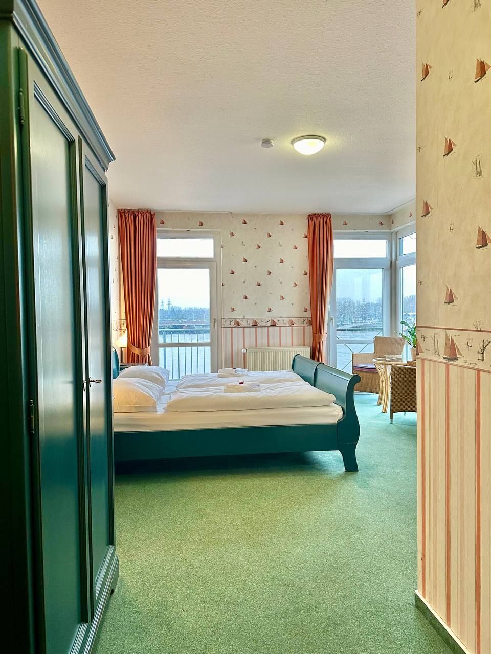 A hotel room with a large bed, two windows with curtains, a radiator, and a view of the city.