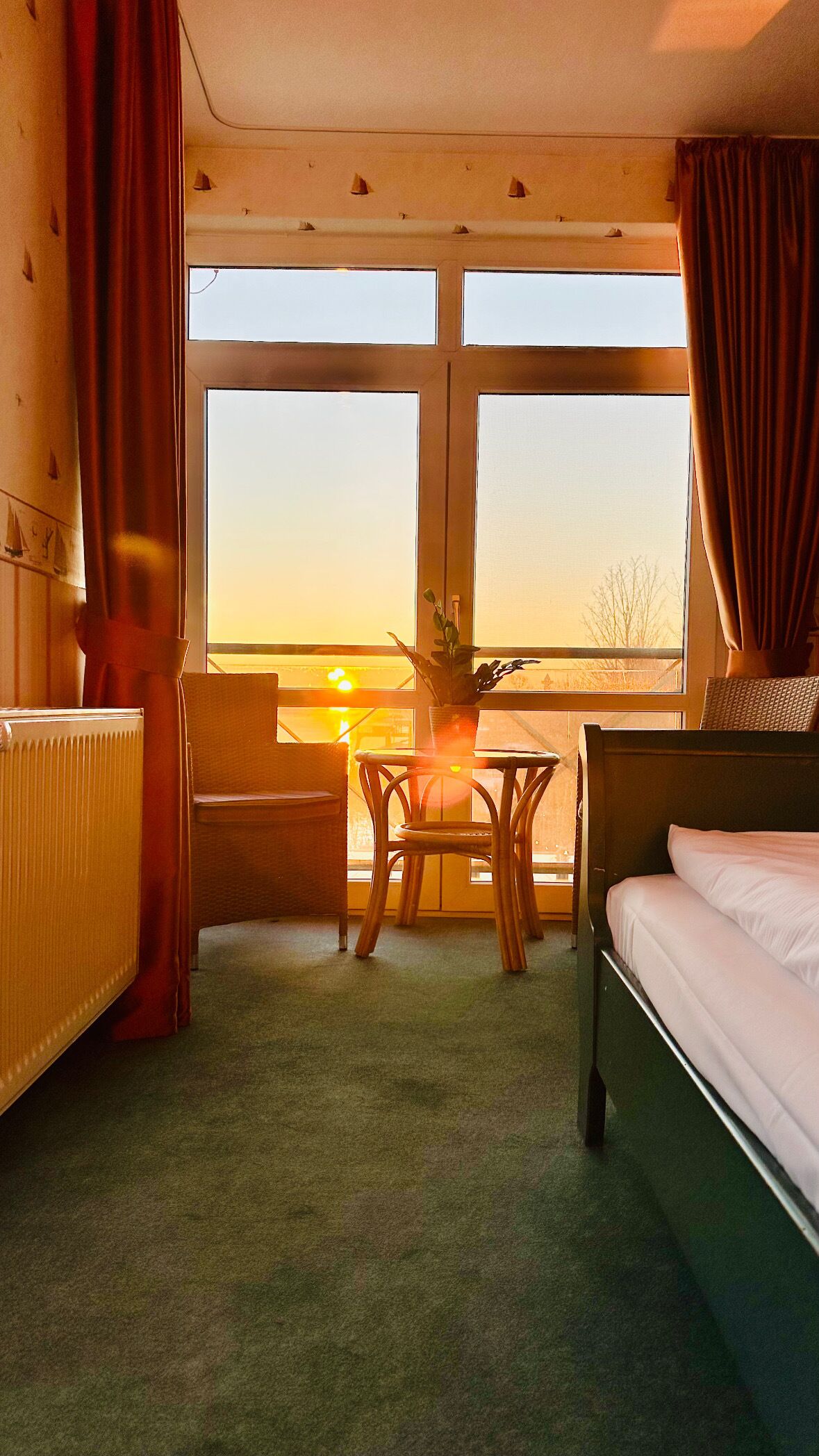 A hotel room with a bed, a chair, a small table, and a view of the sunset through large windows.