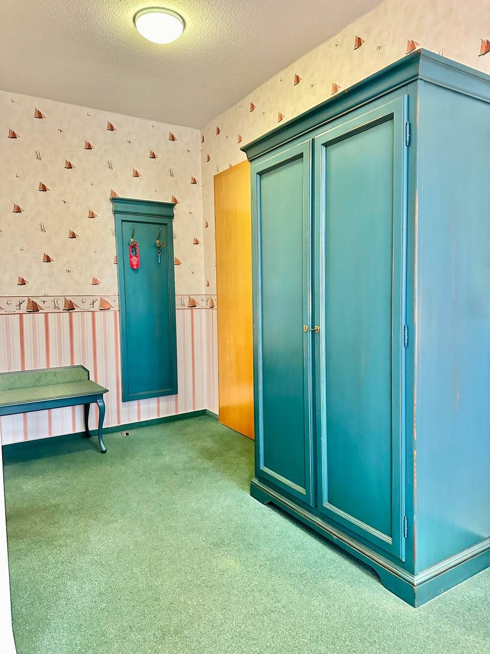 Una habitación con un armario grande color aguamarina, un banco y una puerta con un picaporte rojo de emergencia.