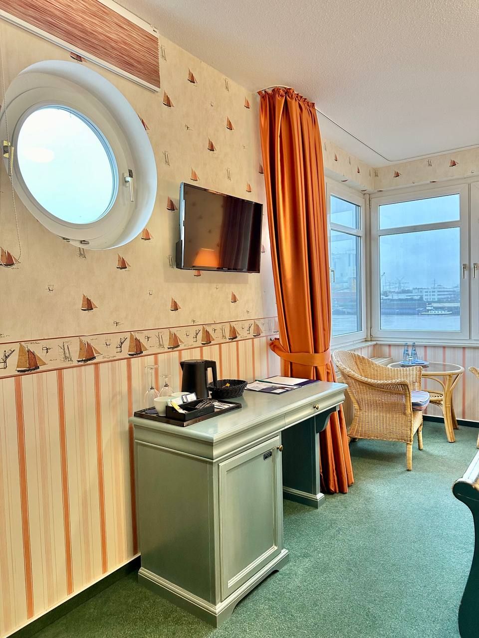 A room with a round window, a flat-screen TV, a desk with a coffee maker, and a chair.