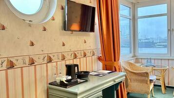 A room with a round window, a flat-screen TV, a desk with a coffee maker, and a chair.
