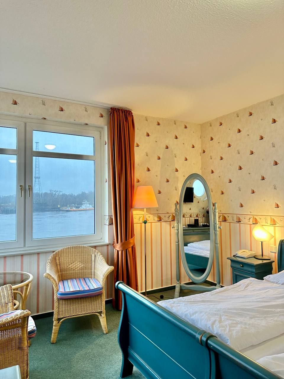 A hotel room with a bed, a chair, a desk, a mirror, and a window overlooking a body of water.