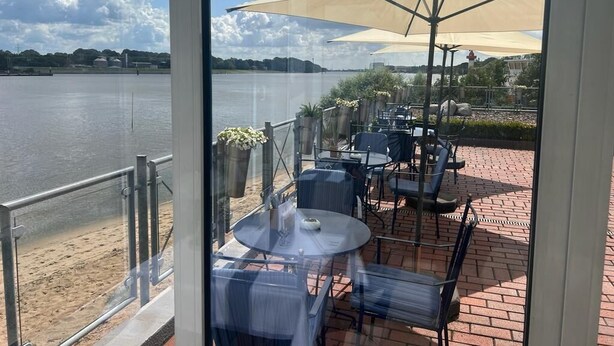 An outdoor patio with tables and chairs, a large umbrella, and a view of a body of water. - Gästehaus Weserblick (Berne)