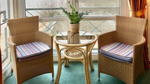 A room with two wicker chairs, a small round table with a potted plant, and a view of a body of water through large windows. - Gästehaus Weserblick (Berne)