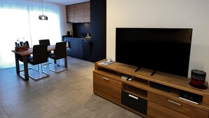 Un salon moderne avec un grand téléviseur à écran plat posé sur un meuble en bois, un coin repas avec une table et des chaises, et une cuisine en arrière-plan.
