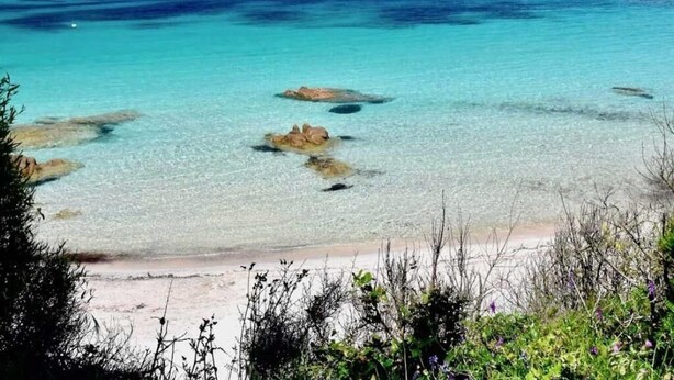 A beach with clear turquoise water, rocky outcrops, and distant mountains.