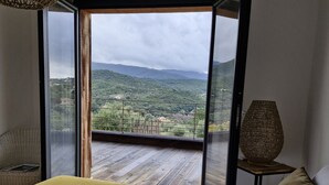 A bedroom with a large bed, a view of a mountainous landscape through an open door, and a wicker lamp on a side table.