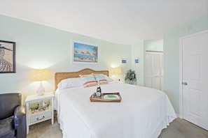 A neatly arranged bedroom with a bed, bedside lamps, a nightstand, a chair, and a door. - Tropical Condo on River | Pools, Spas, & Tennis (Marco Island)