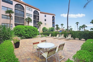 A courtyard with a round table and chairs, surrounded by palm trees and a building with large windows. - Tropical Condo on River | Pools, Spas, & Tennis (Marco Island)