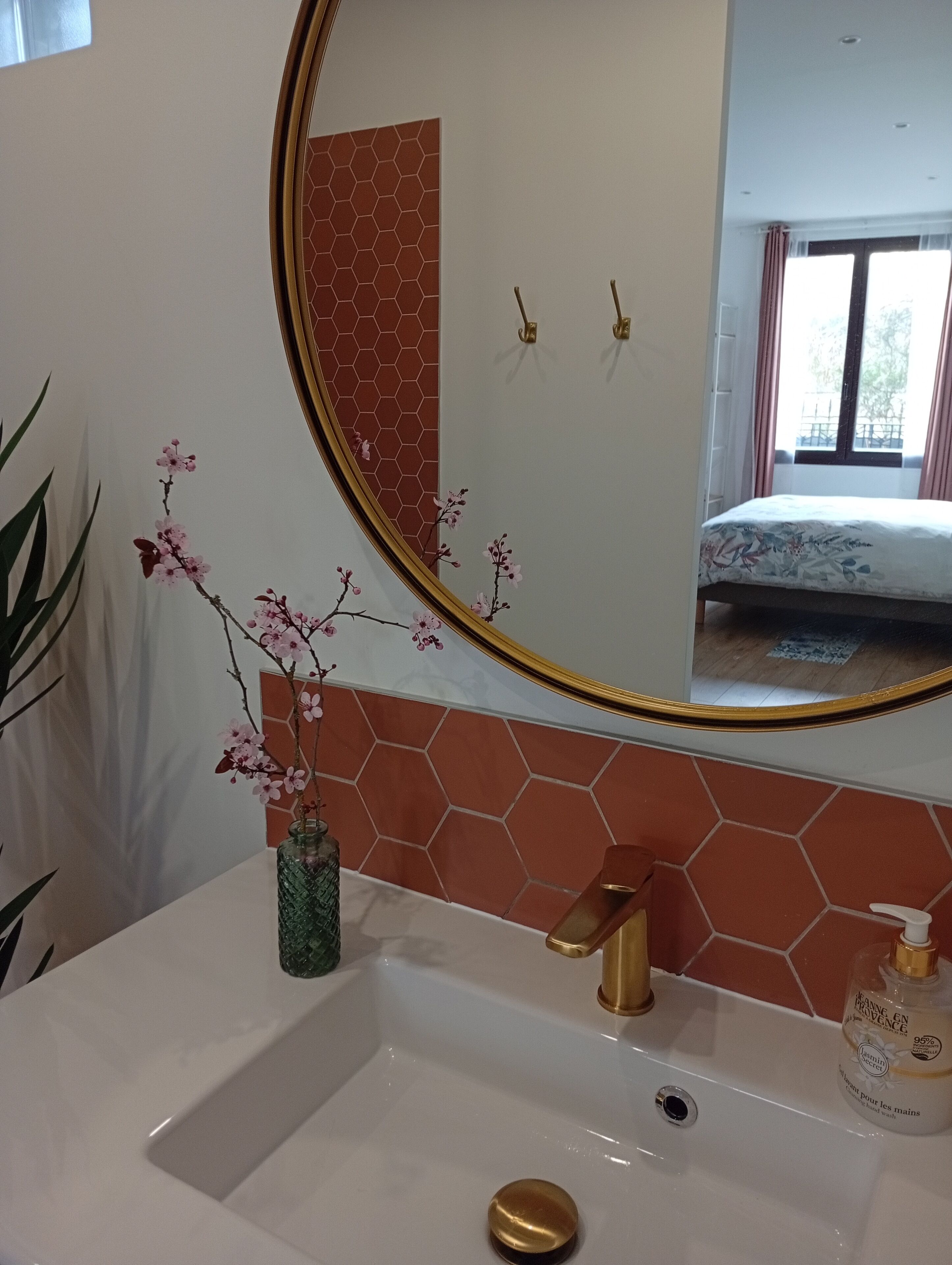 Une salle de bain avec un lavabo blanc, un robinet doré et un miroir avec un cadre doré. On peut voir un vase avec des fleurs roses sur le lavabo, et un flacon de savon sur le côté droit.