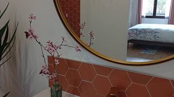 Une salle de bain avec un lavabo blanc, un robinet doré et un miroir avec un cadre doré. On peut voir un vase avec des fleurs roses sur le lavabo, et un flacon de savon sur le côté droit.