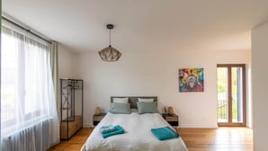 Une chambre à coucher avec un grand lit, un luminaire suspendu, un tableau et une fenêtre ornée de rideaux légers.