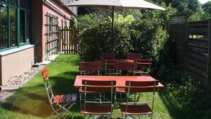 Un espacio de comedor al aire libre con mesas y sillas rojas, un gran paraguas y un edificio de ladrillo al fondo.