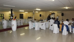A banquet hall with round tables covered in white tablecloths, chairs with white covers, and floral centerpieces. - SHI Hotel (Hukuntsi)