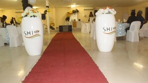 A hotel lobby with a red carpet leading to a reception area, white chairs, and two large vases with the hotel's logo. - SHI Hotel (Hukuntsi)