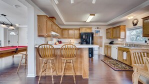 A spacious kitchen with wooden cabinets, a central island with bar stools, a built-in refrigerator, and a pool table in the adjacent room.