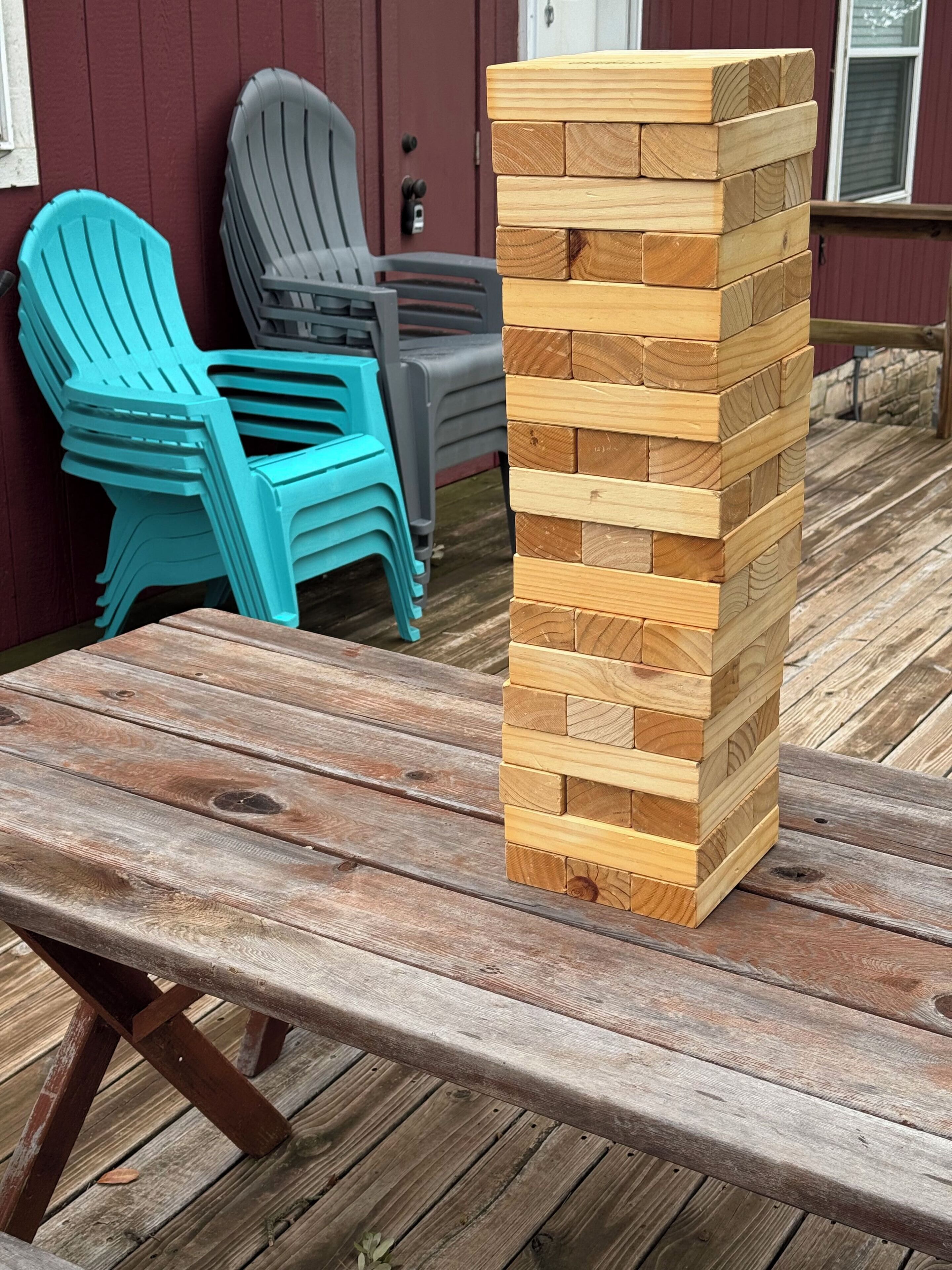 Une tour faite de blocs de bois, sur une terrasse en bois, avec des chaises en plastique et une porte visible au fond.