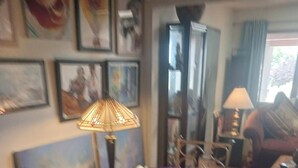 A dining area with a round table covered by a purple tablecloth, surrounded by metal chairs. There are framed artworks on the walls, a tall floor lamp, and a window with curtains. - Randy's Roost -- Private unit within an artists home/studio and gallery. (Santa Fe)