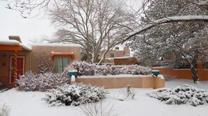 Una zona residencial cubierta de nieve, con un edificio color crema, una puerta roja y arbustos cargados de nieve.