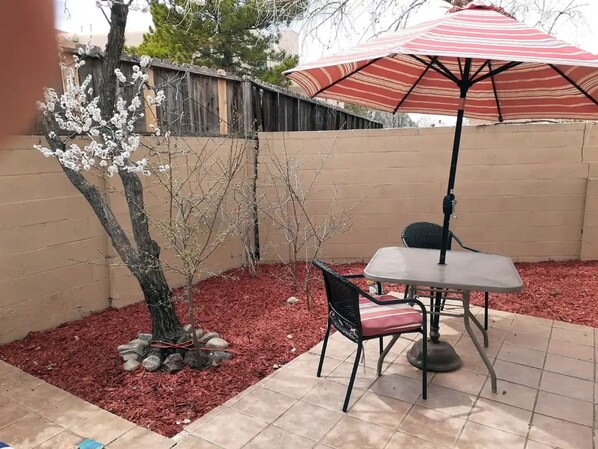 Un patio con mesa y sillas, una sombrilla a rayas y un árbol con flores blancas.