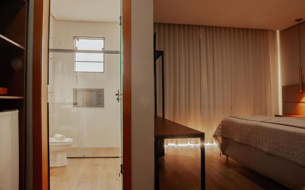 A modern hotel room with a glass door leading to a bathroom, a bed with a headboard, and a bedside table.