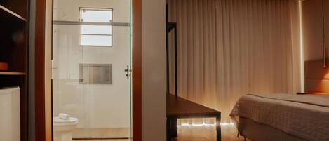 Une chambre d’hôtel moderne dotée d’une porte en verre donnant sur la salle de bain, d’un lit avec une tête de lit et d’une table de chevet.