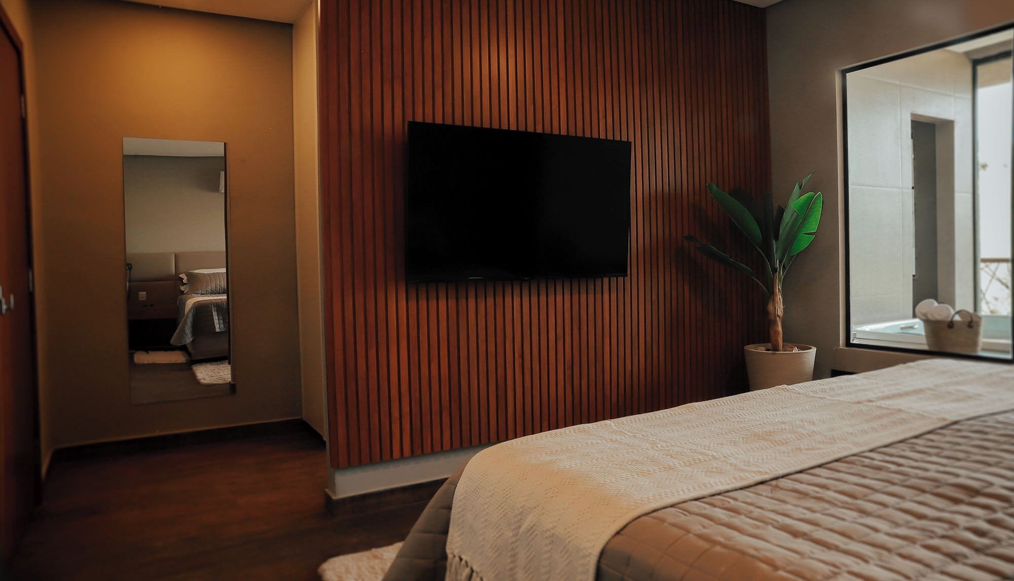 A modern hotel room with a large bed, a flat-screen TV mounted on the wall, a potted plant, and a large window.