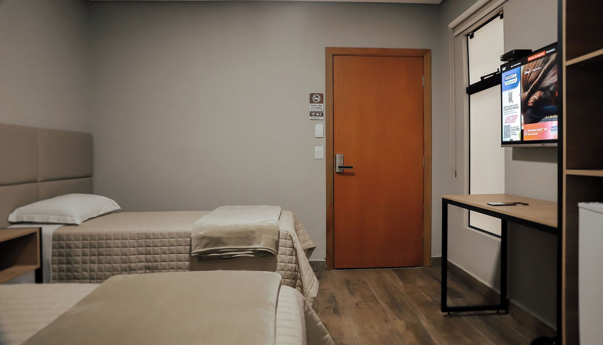 Habitació d'hotel amb dos llits, una porta de fusta, un taulell petit i un televisor fixat a la paret.