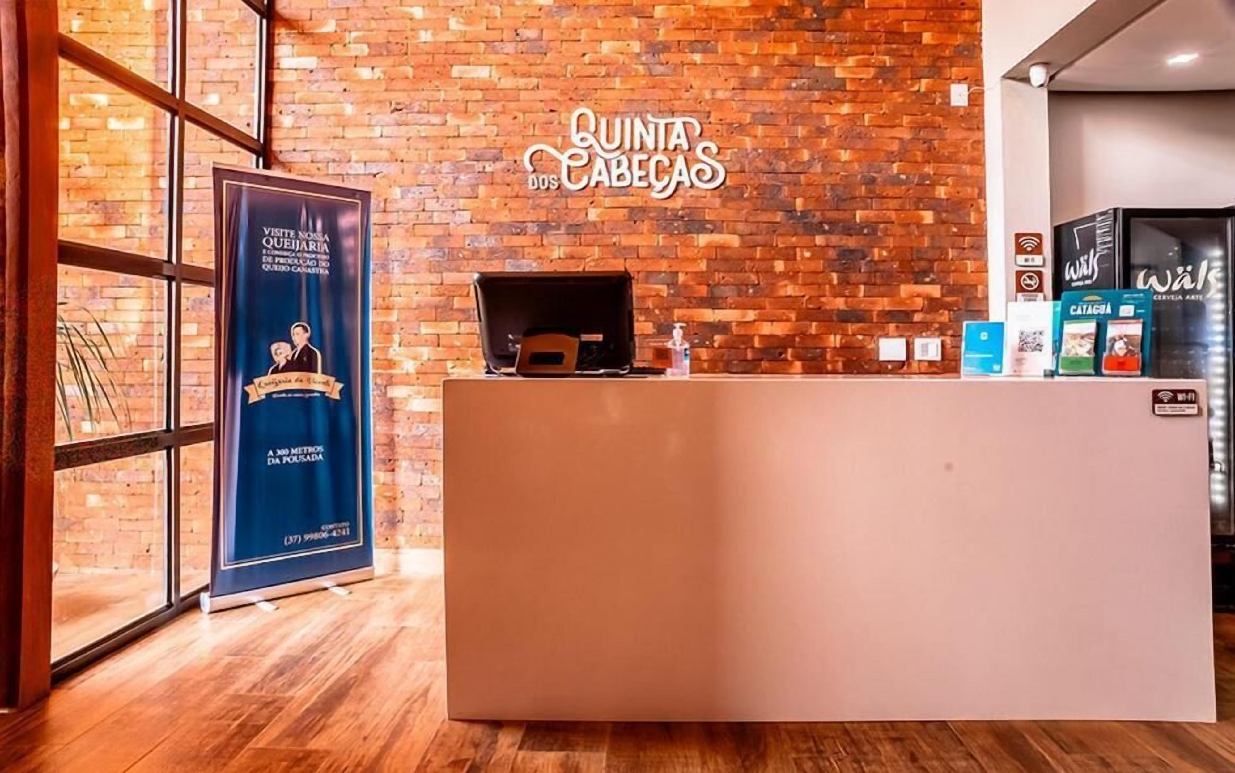 Balcão de recepção com parede de tijolos ao fundo, um cartaz anunciando uma visita à Queijaria e um símbolo de Wi-Fi indicando acesso à internet.