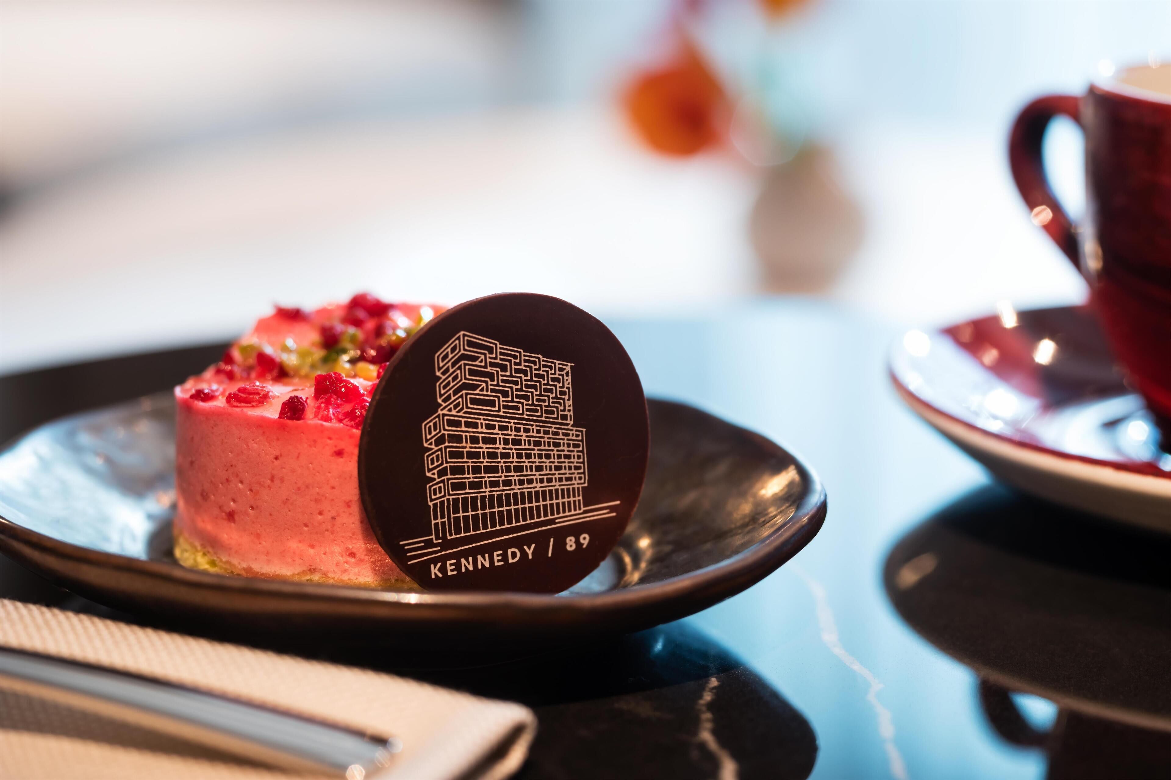 Una rebanada de pastel con un plato decorativo que muestra una ilustración de un edificio y el texto "KENNEDY / 89".