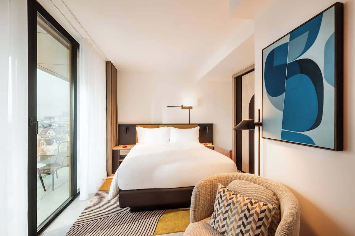 Habitación de hotel moderna con una cama grande, vistas a través de un ventanal, una silla y un cuadro abstracto de grandes dimensiones en la pared.