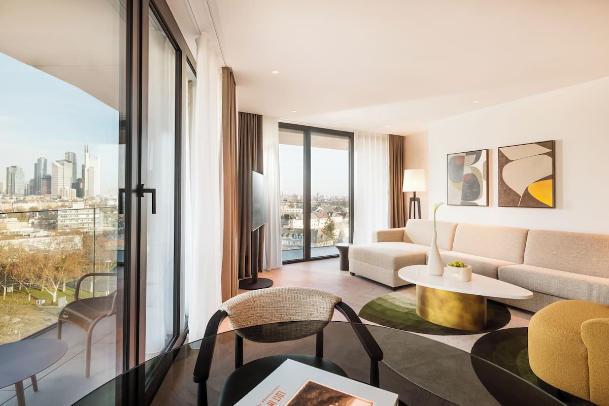 Una habitación de hotel moderna con un ventanal grande que ofrece vistas a la ciudad, un sofá, una mesa de centro y una silla.