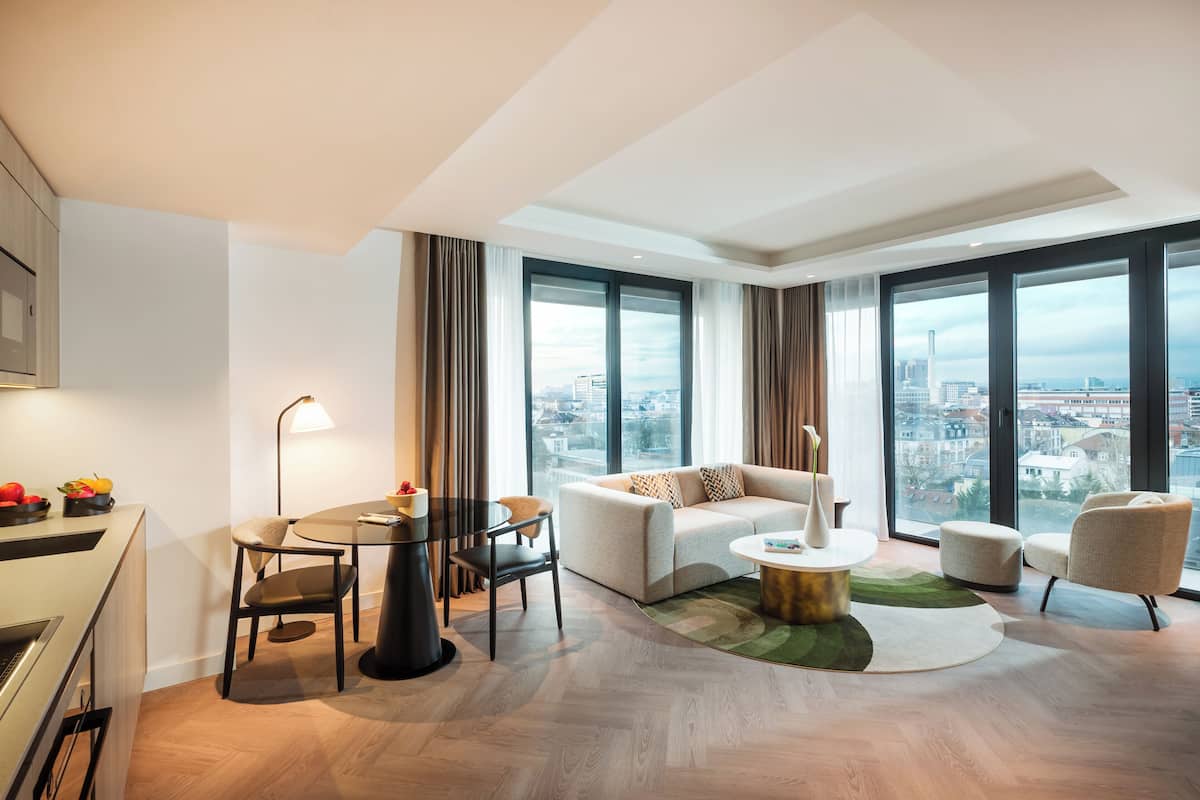 Una sala de estar moderna con un sofá, sillas, una mesa de comedor y ventanas grandes que ofrecen vistas a la ciudad.
