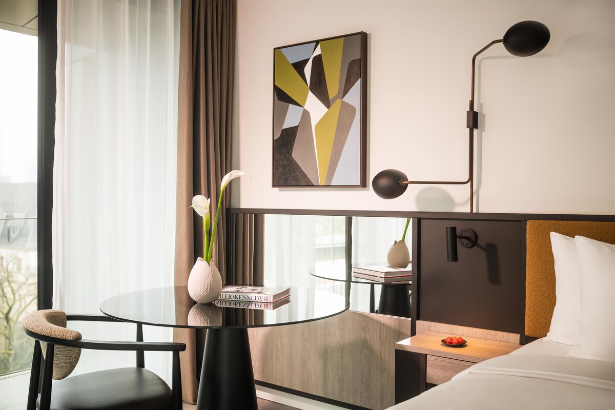 A modern hotel room with a glass table, a comfortable chair, a bed with a headboard, a bedside table with a vase, and a wall-mounted lamp.