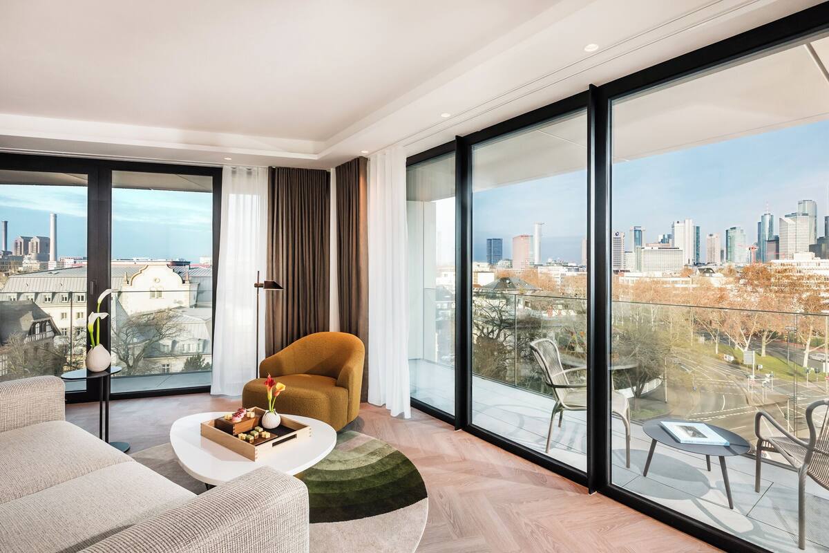 Un salón moderno con sofá, mesa de centro y una silla. Se ve un ventanal con vistas a la ciudad.