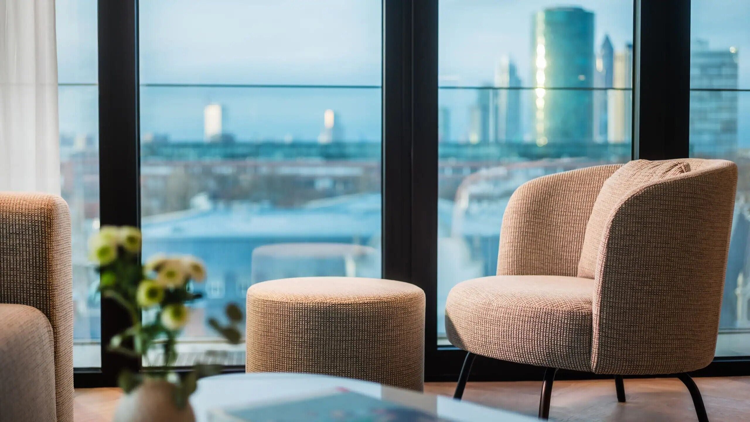 Una habitación moderna con un amplio ventanal que ofrece vistas a la ciudad, un sillón cómodo, un pequeño reposapiés y un jarrón con flores.