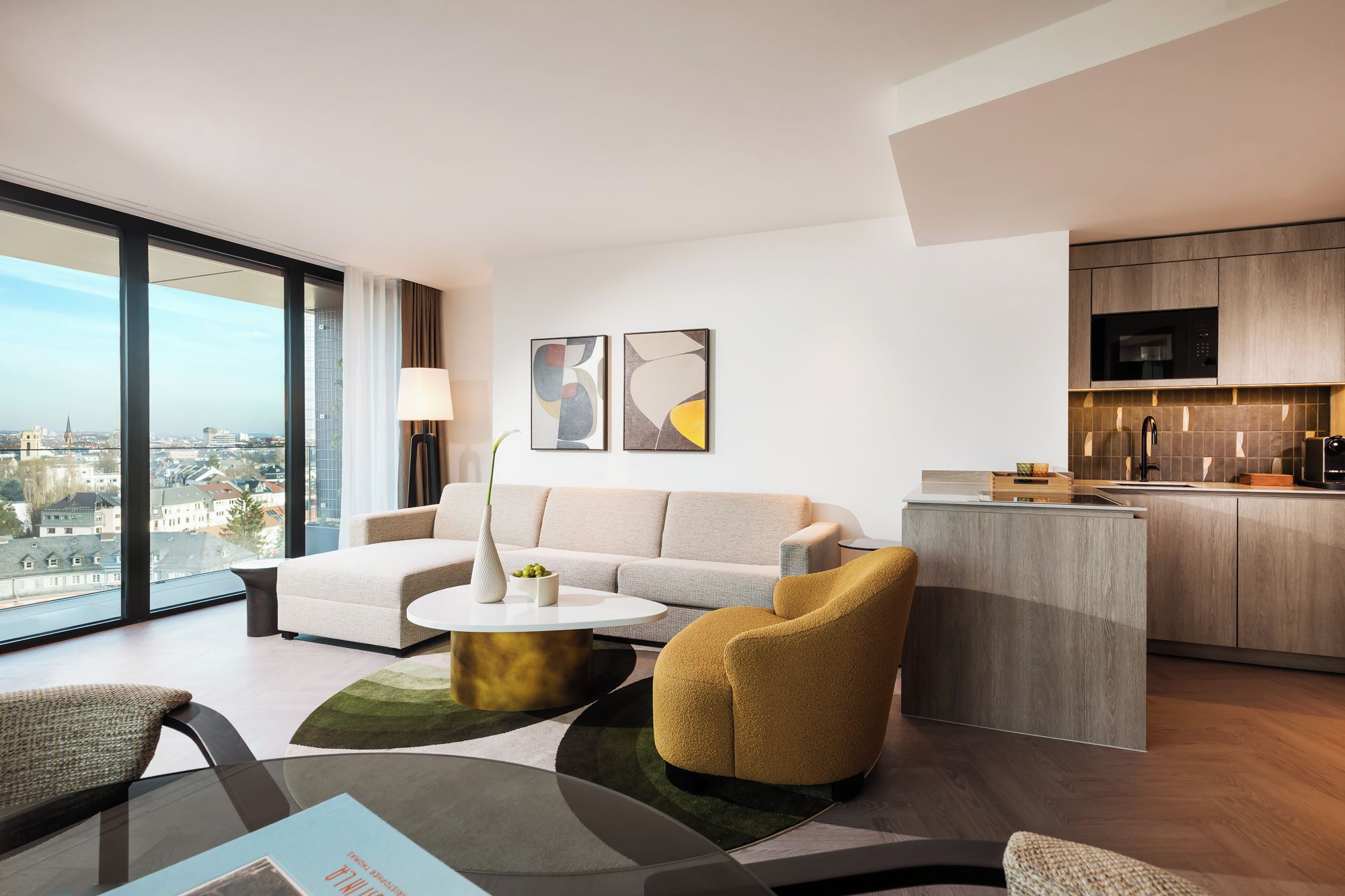 A modern living room with a sofa, armchair, and coffee table, a kitchen with a microwave, and a large window offering a city view.