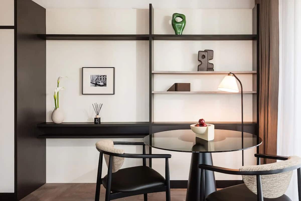 Un comedor moderno con una mesa redonda, dos sillas, una lámpara de pie y una estantería minimalista con objetos decorativos.
