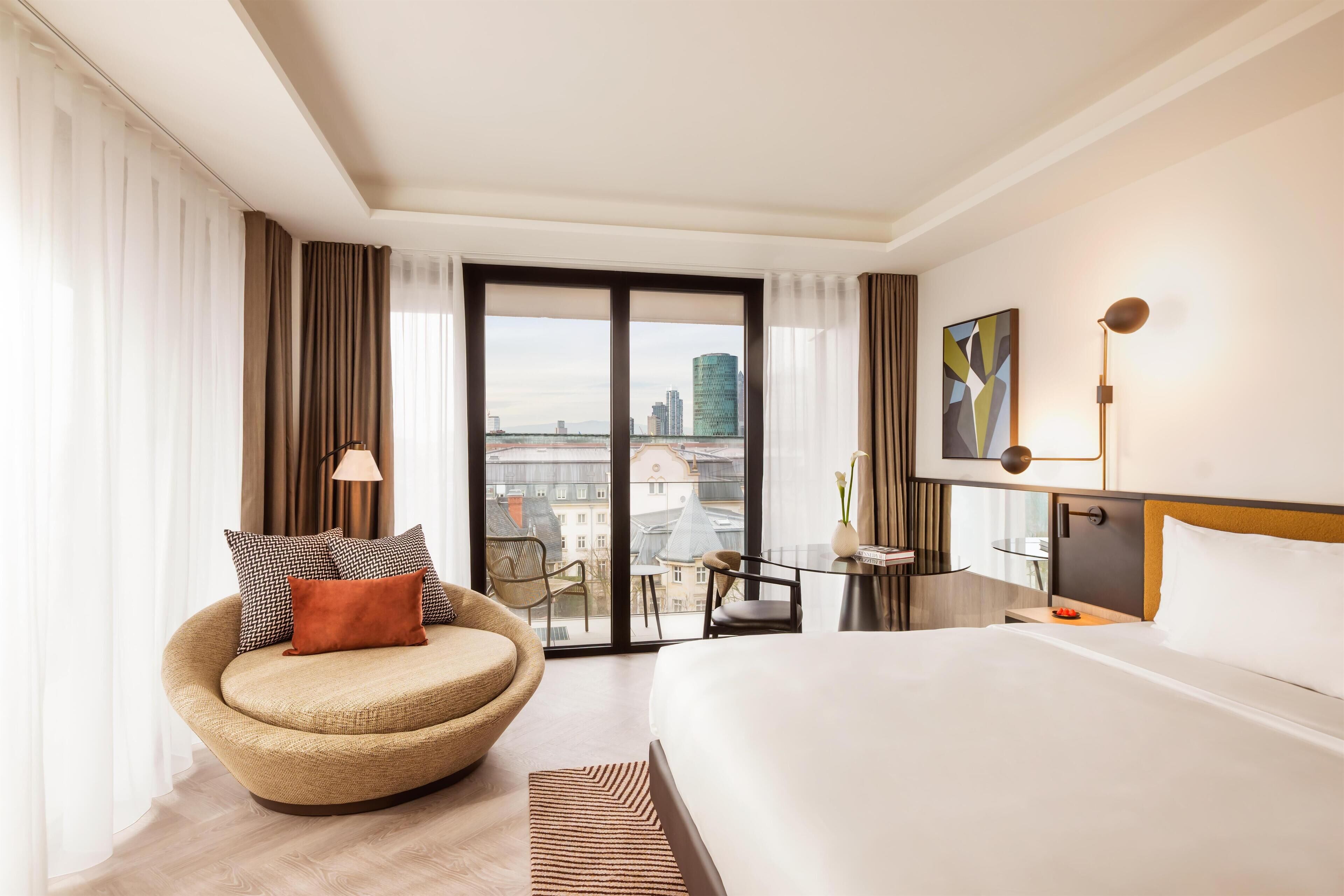 Ett modernt hotellrum med en stor säng, en böjd fåtölj, ett skrivbord med en stol och utsikt över stadsbilden genom fönstret.