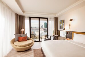 A modern hotel room with a large bed, a curved armchair, a desk with a chair, and a view of the cityscape through the window. - Kennedy 89 Hotel Frankfurt, part of Hyatt (Frankfurt)